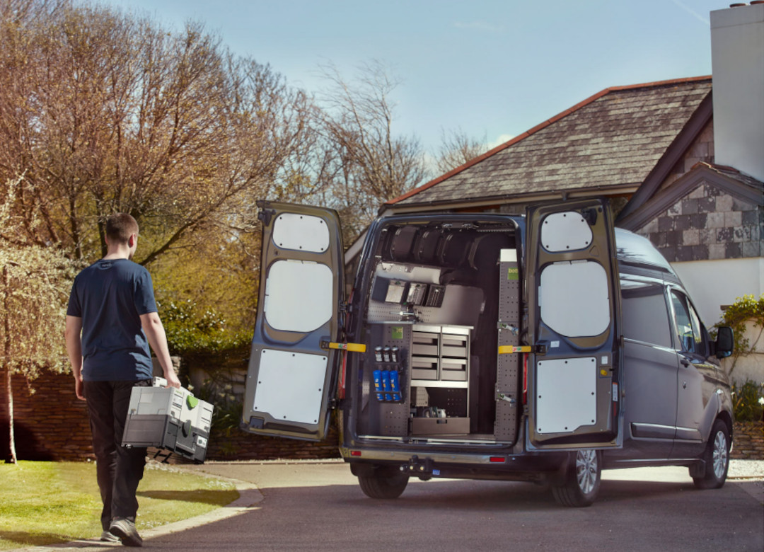 man on house driveway approaching rear of van with open doors, carrying two stacked bott boxes, ready to stack back into van racking